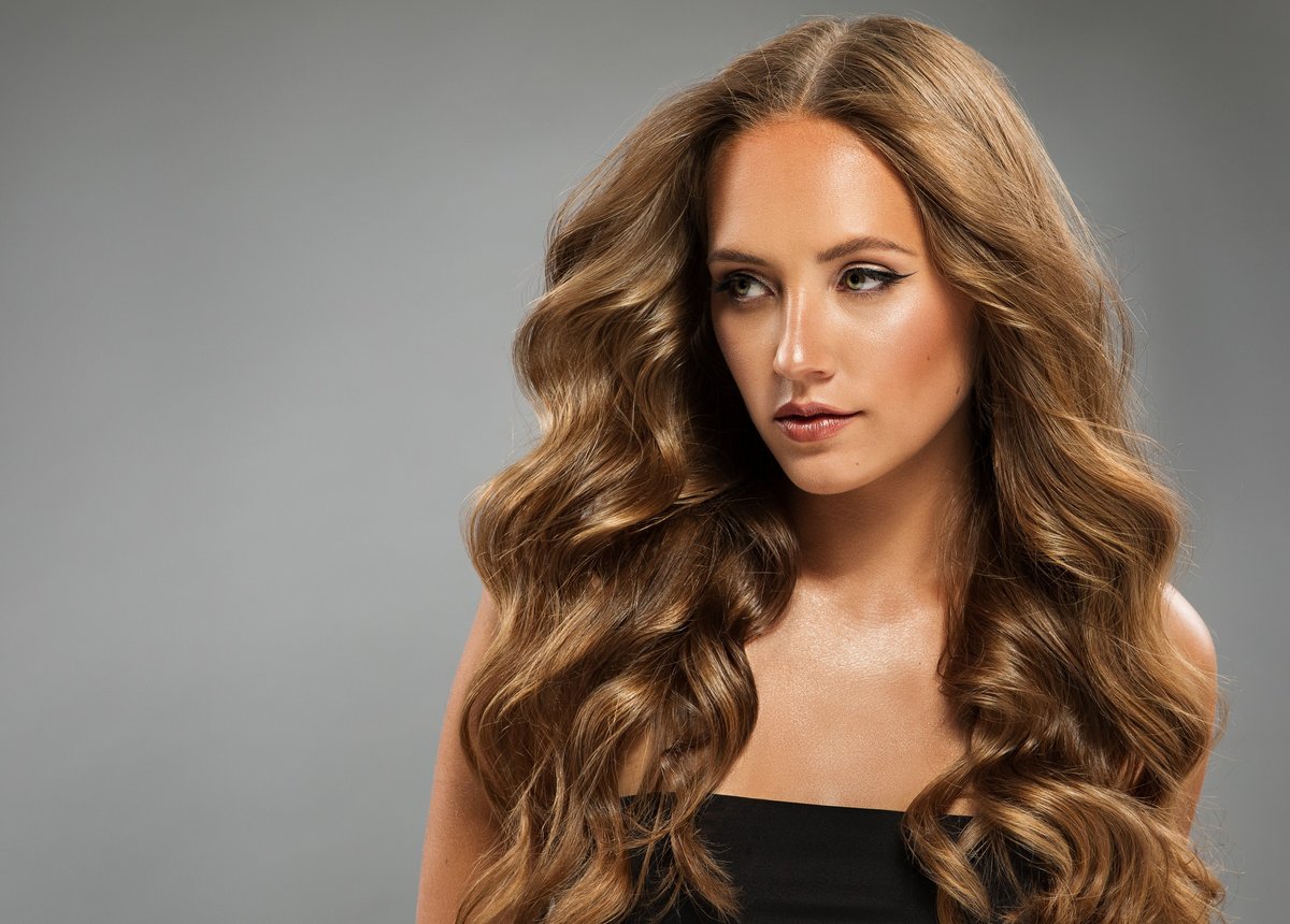 Curly Hair Model. Beautiful Girl with Long Wavy Hairstyle over Gray Background. Fashion Woman with natural Makeup and Volume Hairdo looking away