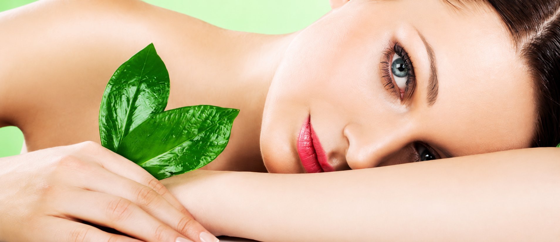 Woman with glowing skin and red lips holding fresh green leaves against soft green background